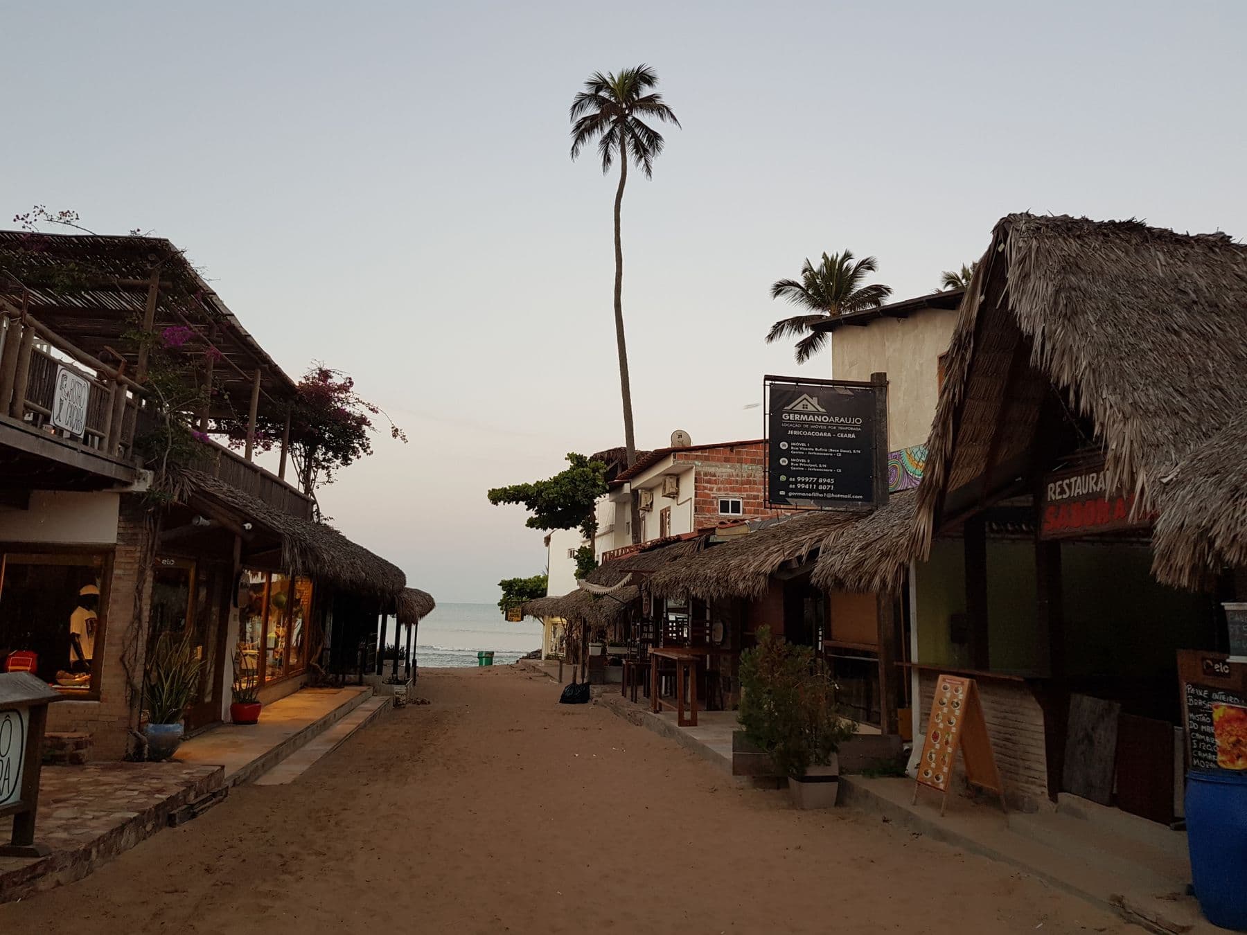 Rua de areia do vilarejo de Jericoacoara ao anoitecer, com pousadas de telhado de palha e um coqueiro alto