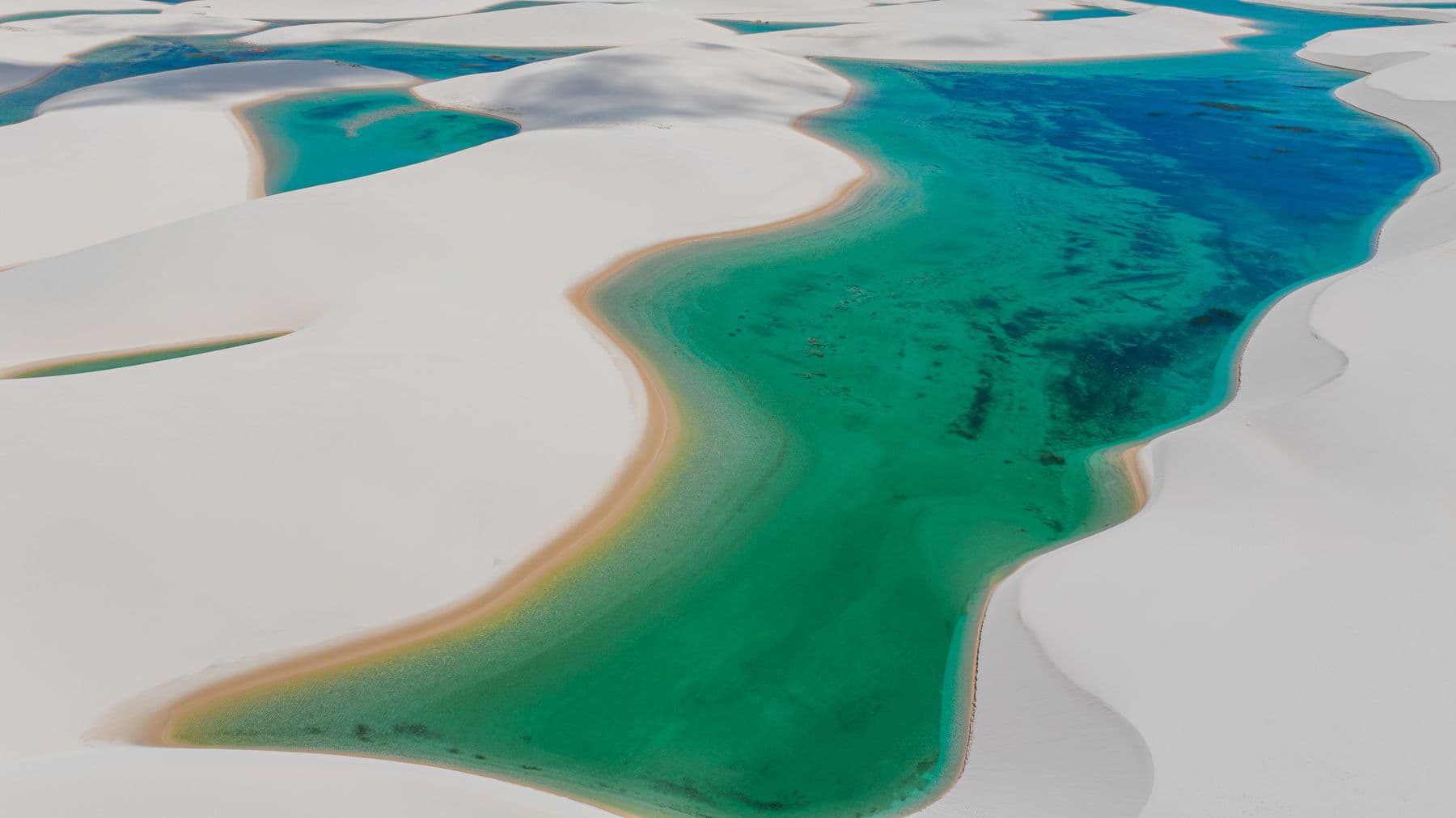 Lagoa de água esmeralda serpenteando entre dunas brancas, vista de cima, nos Lençóis Maranhenses