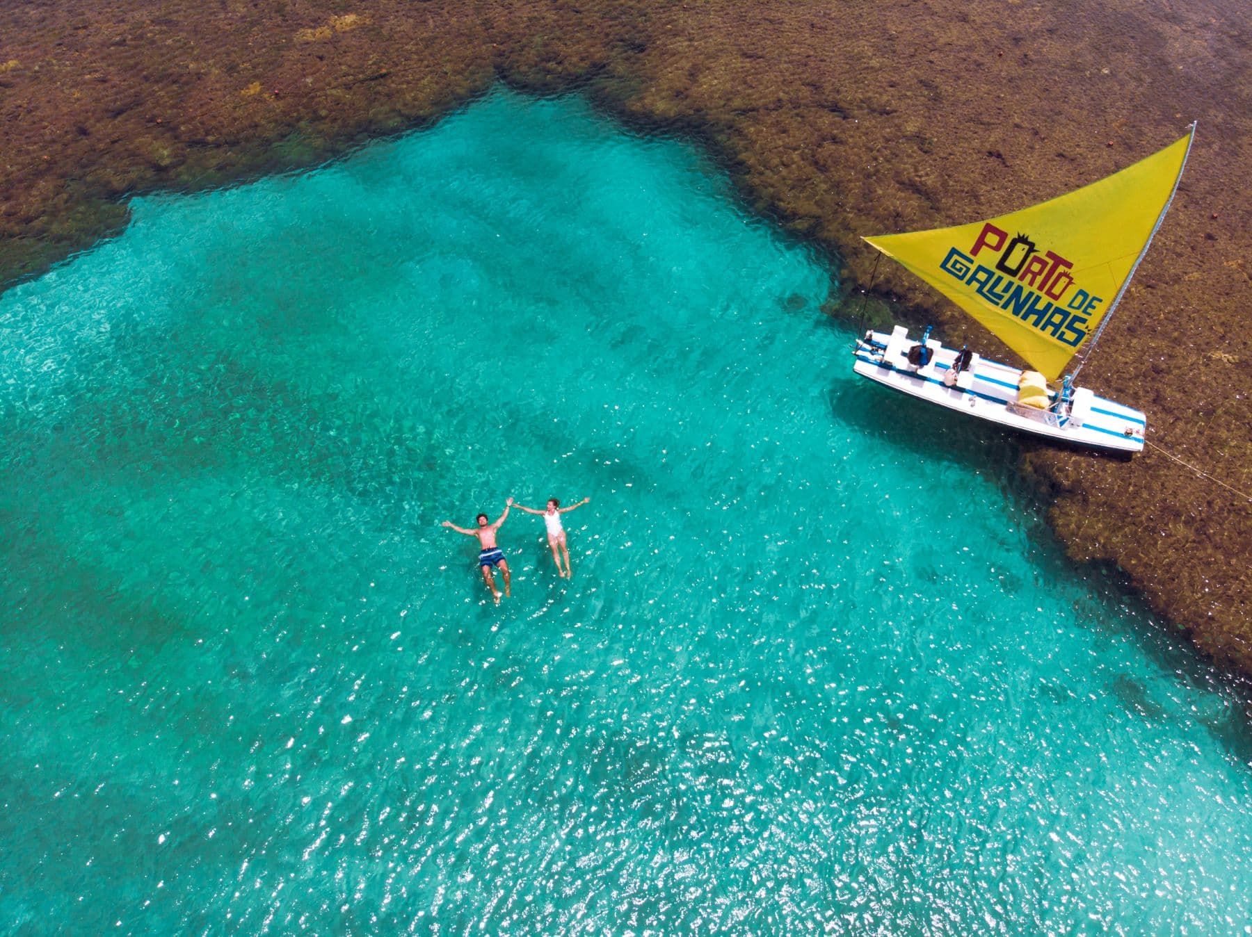 Vista aérea de duas pessoas boiando nas piscinas naturais de água turquesa de Porto de Galinhas, ao lado de uma jangada