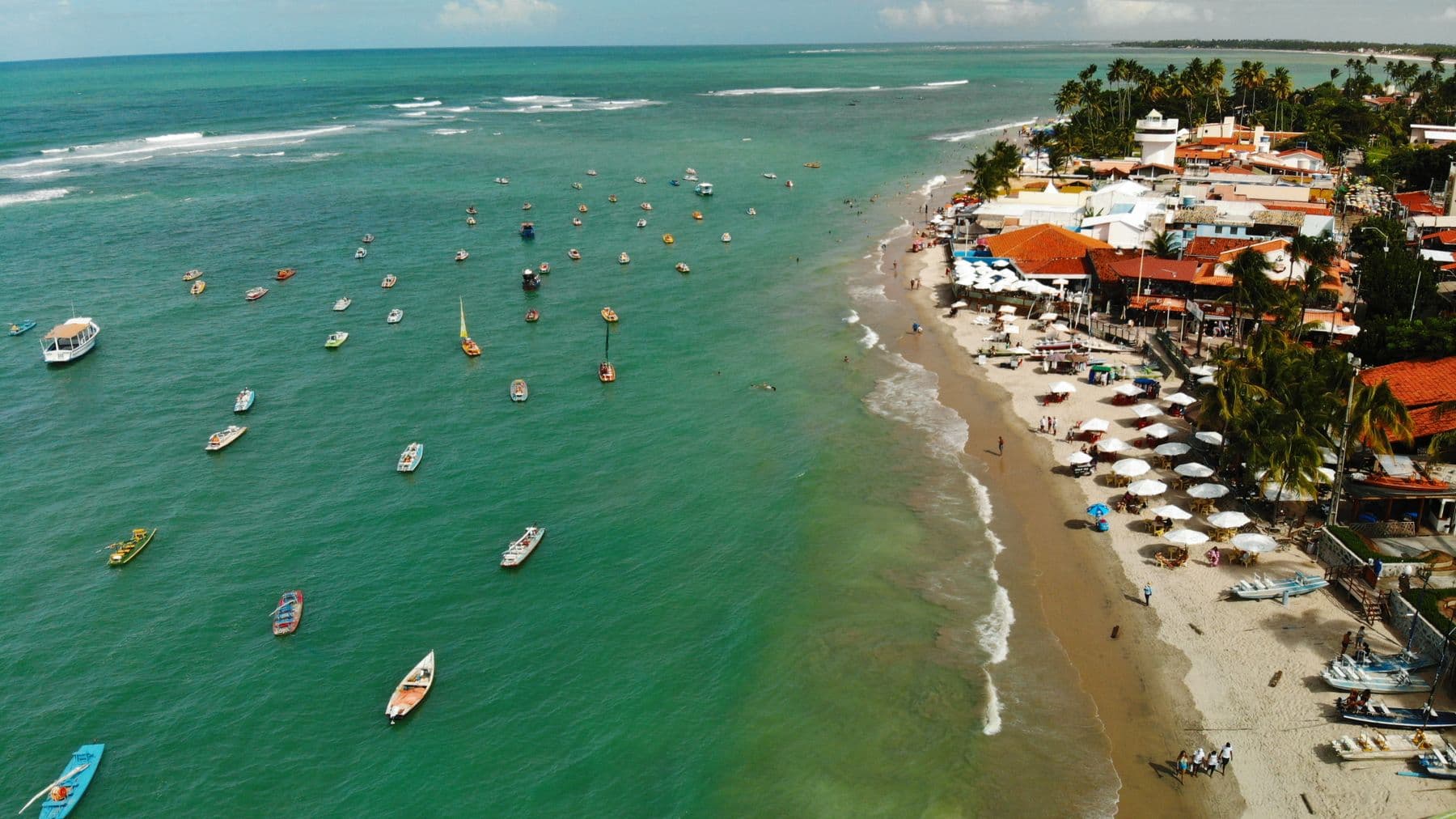 Vista aérea da orla de Porto de Galinhas, com dezenas de jangadas ancoradas no mar esverdeado
